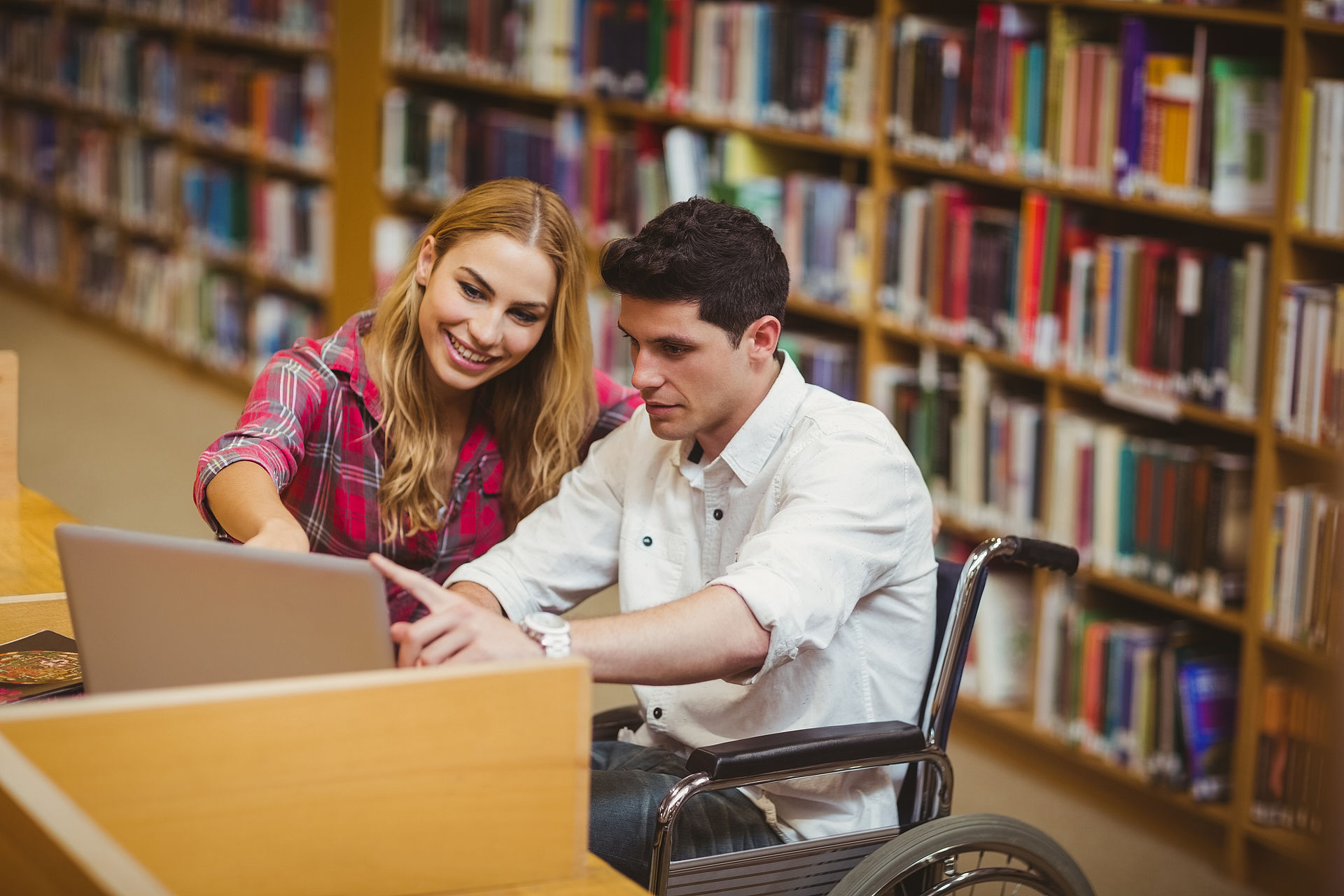 Studentin in der Bibliothek zeigt Studenten im Rollstuhl etwas am Laptop