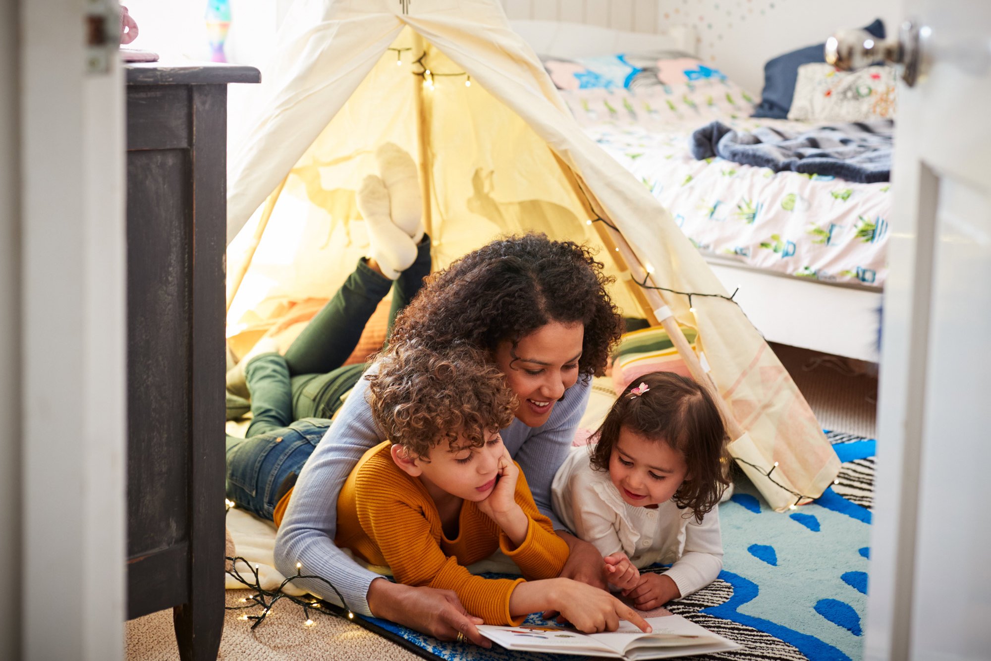 A mother is lying on the floor with her two small children and reading to them. 