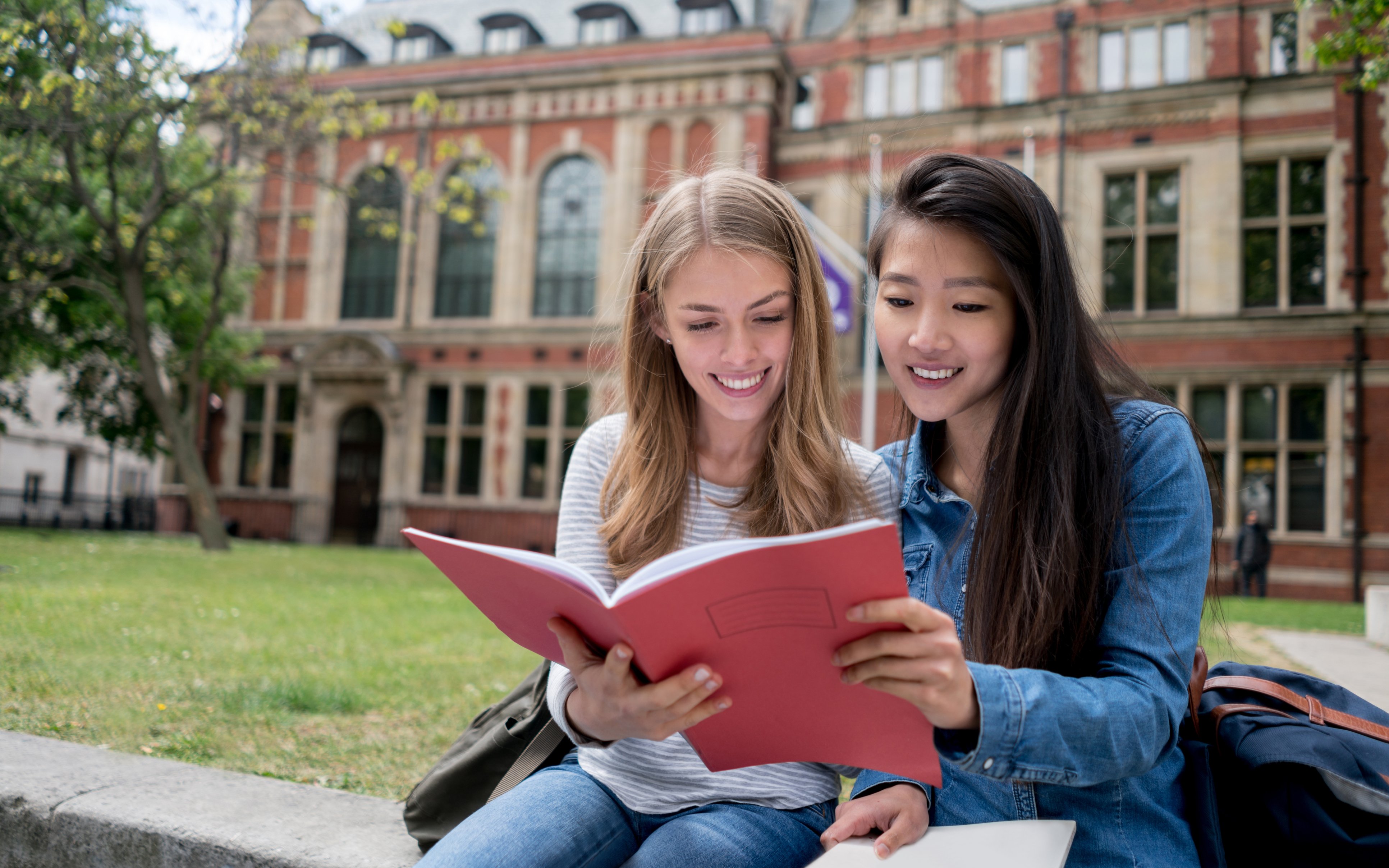 Zwei Studentinnen sitzen vor der Uni und schauen gemeinsam in eine Broschüre o.ä.