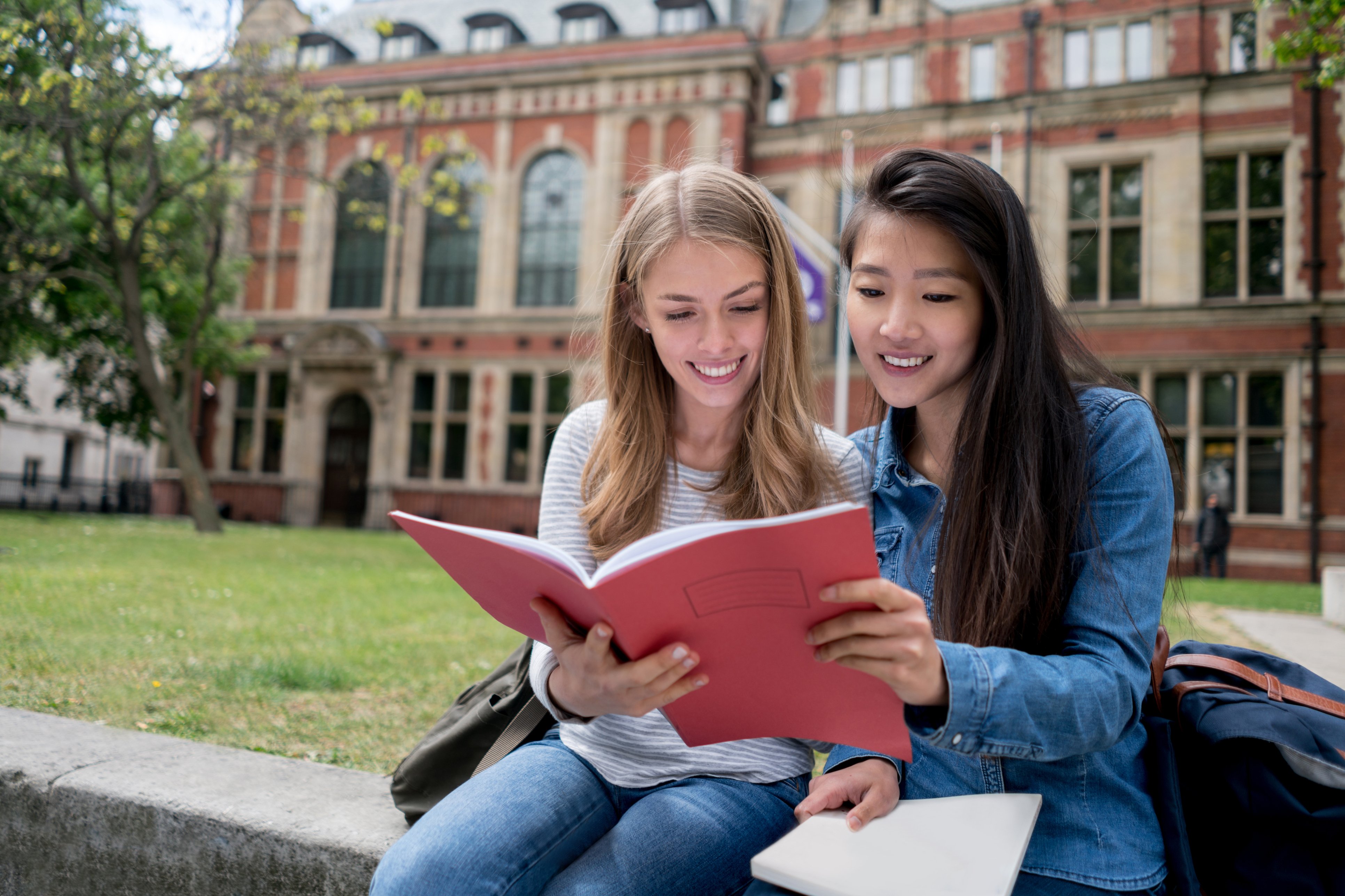 Two female students are sitting in front of the university, looking at a brochure or something similar together.